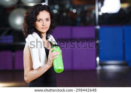 Waist up portrait of fitness woman with curly short hair in sports wear with towel and green bottle on a sports equipment background at the gym