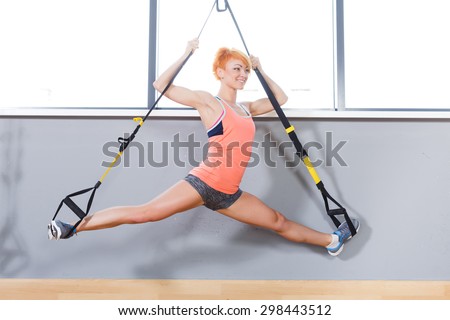 Young attractive redhead woman does splits crossfit stretching with trx fitness straps in the gym