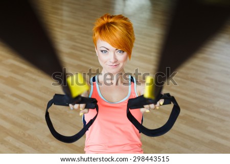 Pretty redhead young fitness girl in shirt doing exercises with TRX in gym