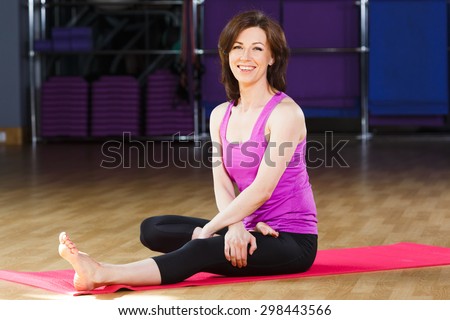Young laughing woman with dark straight short hair wearing on violet shirt and black leggings is sitting on pink yoga mat on a sports equipment background at the gym