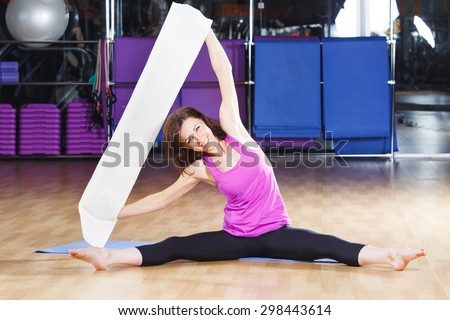 Smiling woman with dark hair wearing on violet shirt and black leggings does exercises on a yoga mate with a white core band on a sports equipment background at the gym