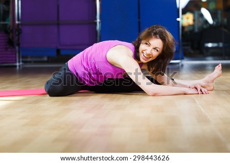 Laughing fitness girl with dark straight hair wearing on violet shirt and black leggings is doing exercises on pink yoga mat on a sports equipment background at the gym