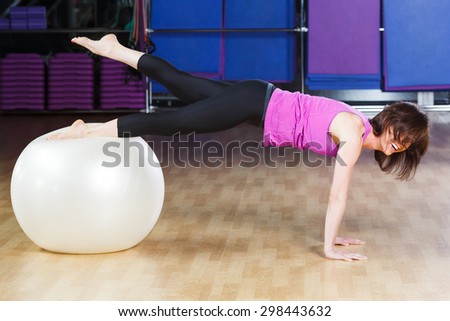 Attractive young fitness woman with dark hair wearing on violet shirt and black leggings does exercises and laughing on a white fitball at the gym