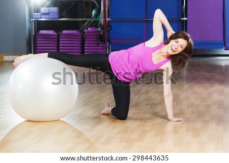 Nice young fitness woman with short dark hair wearing on violet shirt and black leggings does exercises and laughing on a white fitball at the gym