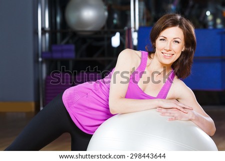 Young, smiling fitness woman with short dark hair wearing on violet shirt does exercises on a white fitball at the gym