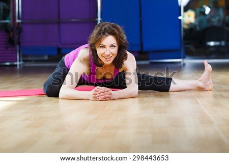 Smiling fitness girl with dark straight short hair wearing on violet shirt and black leggings is doing exercises on pink yoga mat on a sports equipment background at the gym