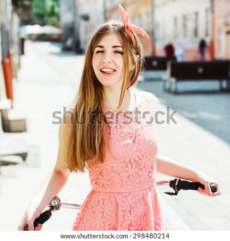 Pretty girl having fun and showing tongue, holding a bicycle handlebar in a sunny day on the street of the old town. Young woman wearing pink head wrap and pink dress with a pattern of flowers.