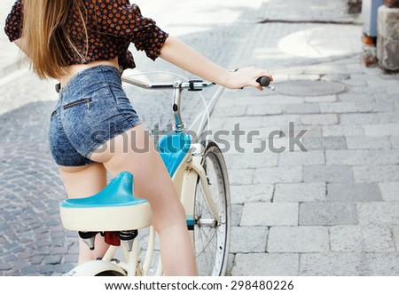 Pretty girl smiling and holding a bicycle handlebar in a sunny day on the street of the old town. Young woman with blond-brown long hair, wearing pink head wrap and pink dress with pattern of flowers