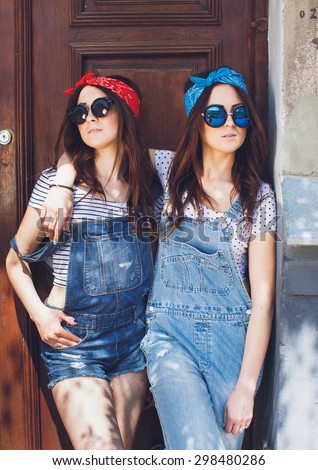 Attractive young brunette twins girls, in stylish sunglasses, hugging. Wearing denim overalls bright bandanas, posing in front of old doors. Outdoors. Summer day.