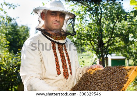 Serious apiarist is posing with frame of honeycomb with a lot of working bees, in the yard, waist up