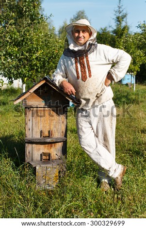 Apiarist in protective hat near the wooden beehive with roof is standing in the bee-garden with multicolor wooden beehives on the background, close up