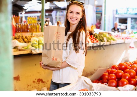 Attractive smiling girl, with paper bag of products is standing near the counter with tomatoes, during the shopping at fruit vegetable market, waist up