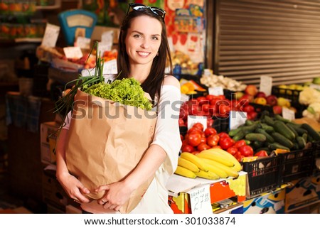 Smiling girl, with dark long hair, is holding a paper shopping bag full of groceries, at fruit vegetable market, on counter with vegetables and fruits background, waist up