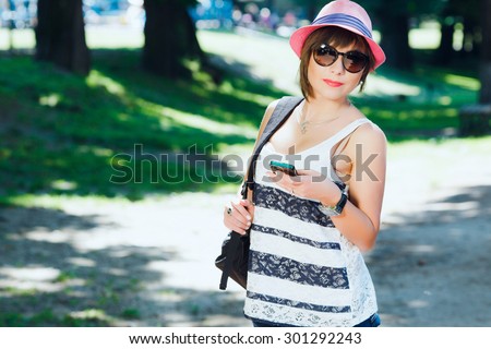 Close up portrait of cute young woman, with smart phone and black backpack on shoulder. Looking at camera. Wearing sunglasses, stylish pink hat and striped shirt. On sunny day, park alley background.