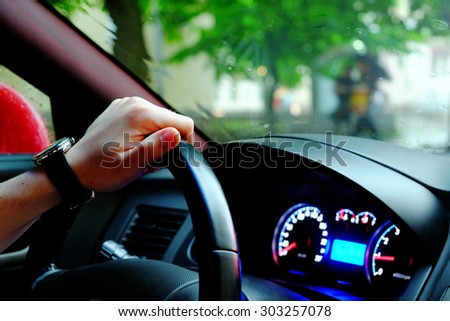 Driver\'s hand on a steering wheel of a car, street with green trees and incidental people on a background, close-up