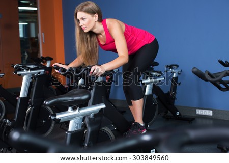 Beautiful brunette woman, wearing in sportswear, doing indoor biking exercise, on blue wall background, full body
