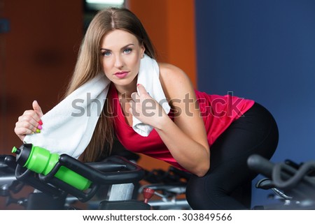 Beautiful brunette woman is sitting on exercise bike with bottle of water and white towel, in the gym, waist up