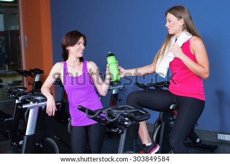 Two attractive young women in sports clothing exercising on gym bicycles. Girl in a pink shirt gives a bottle of water to girl in violet shirt, waist up