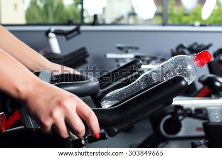 Cropped image of woman\'s hands holding exercise bike, with bottle of water on it, on the gym, with window on a background, close up