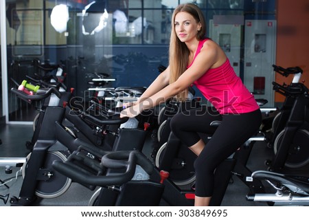 Beautiful athletic woman, wearing in shirt and leggings, posing on exercise bike in gym, full body
