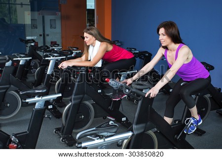 Two beautiful brunette women, wearing on shirts and leggings, doing indoor biking exercise, full body