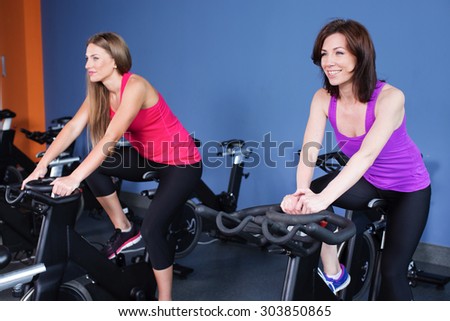 Two beautiful brunette women, wearing in sportswear, doing indoor biking exercise, on blue wall background, full body