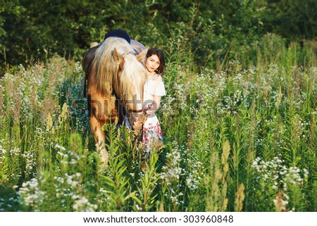 Charming brunette girl, wearing in white dress is holding brown and white horse on the bridle, in the green meadow, full body