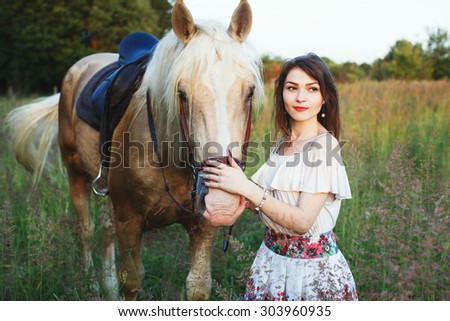 Brunette woman in white short dress, standing with beautiful brown strong and muscular horse in green summer field, waist up