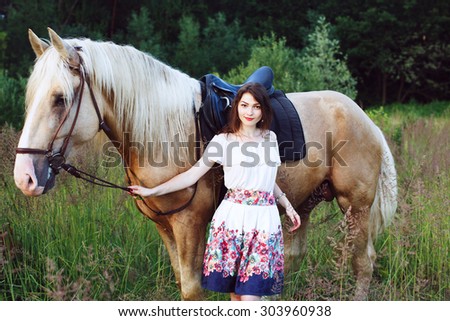 Young pretty brunette woman, wearing on white short dress, is posing with beautiful brown horse, in summer field