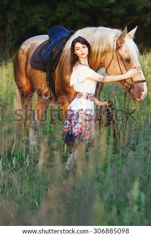 The image of a young brunette woman, wearing in white dress which is holding brown horse on the bridle, in the green meadow, full body