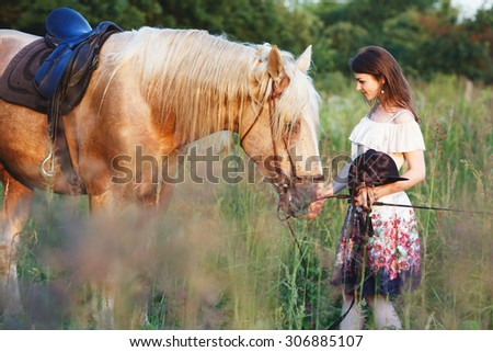 Charming girl in white dress with pattern, holding a brown and white horse on the bridle in green summer field, full body