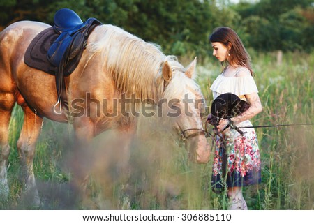 Charming girl in white dress with pattern, standing with brown and white strong horse with saddle, and holding black hat in her hands, in green summer field, full body