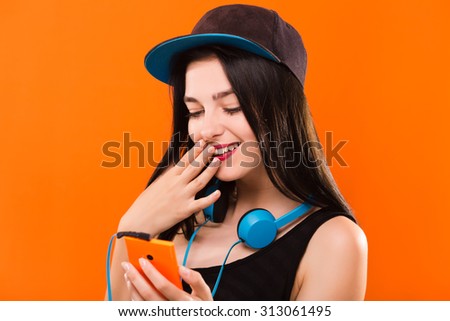 Smiling pretty girl, with blue headphones on her neck, wearing in black blouse and cap, holding smart phone in her hand - isolated on orange background, in studio, waist up