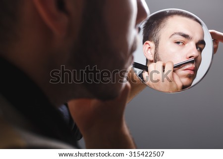 Young brutal man, wearing in black shirt, holding mirror and shaving with razor, on gray background, in studio, close up