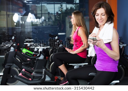Two young fitness women, wearing in shirt and leggings, are sitting on a exercise bikes. Woman in violet shirt is holding smart phone in her hand, waist up