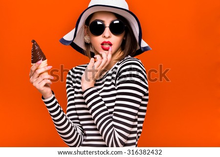 Lovely young girl, wearing in striped blouse, black sunglasses and white hat, is posing with plastic ice cream in her hand, on orange background, in studio, waist up