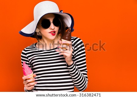 Pretty brunette girl, wearing in striped blouse, black sunglasses and white hat, is posing with two ice cream, on orange background, in studio, waist up