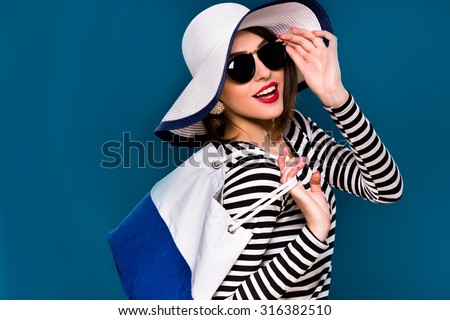 Cute smiling woman, with dark hair, wearing in striped blouse, black sunglasses and white hat, is posing with white and blue bag, on blue background, in studio, waist up