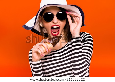 Funny young girl, wearing in striped blouse, black sunglasses and white hat, is holding plastic ice cream in her hand, like she is going to eat it, on orange background, in studio, waist up
