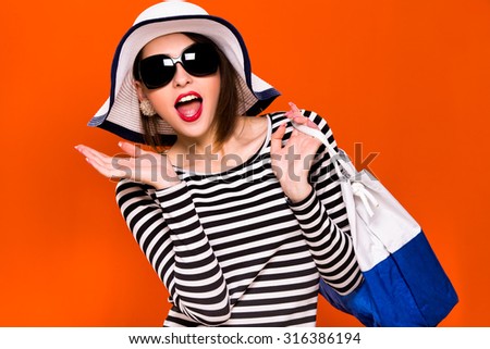 Young happy woman, with dark hair, wearing in striped blouse, black sunglasses and white hat, is standing with white and blue bag, on orange background, in studio, waist up