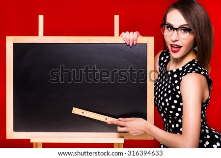 Charming surprised girl, with dark hair, wearing in black with white dots dress and glasses, is showing something on the blackboard with a ruler, on a red background, waist-up