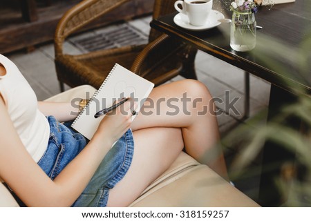 Slim girl, wearing on blue shorts and white shirt, sitting on beige leather sofa and drawing on her notebook, vase with flowers and cup of coffee standing, close up. Point of view