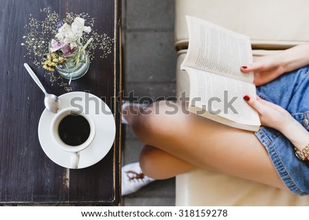 Young woman, wearing on blue shorts, sitting on beige leather sofa and holding a book, cup of coffee and vase with flowers on the dark wooden table, on cafe, close up. Point of view
