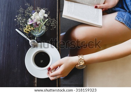 Young girl, wearing on blue shorts, sitting on beige leather sofa, reading a book and holding a cup of coffee, vase with flowers  standing on the dark wooden table, on cafe, close up. Point of view