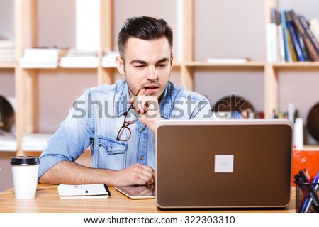 Portrait of handsome man with glasses, wearing in casual blue shirt, sitting at the table and looking at the laptops screen, on the bookshelves background, waist up