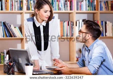Young business woman and man, wearing in casual clothes, are talking, on the bookshelves background, in office, waist up