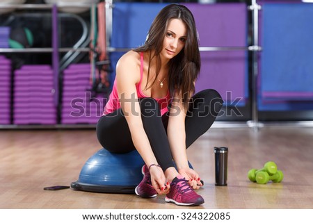 Fitness woman, wearing in shirt, sneakers and black leggings, sitting on a bosu ball and tying her shoe laces. Cup, dumbbells and smart phone lying on the floor, in the gym, full body