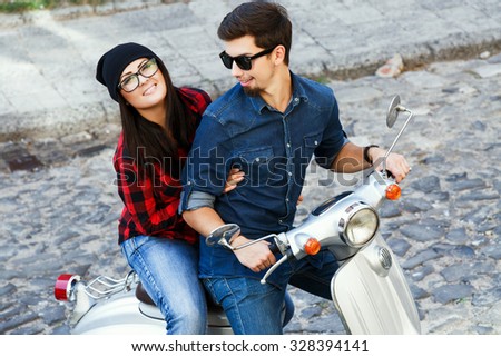 Two beautiful young people, wearing on glasses, shirts and jeans, sitting on a vintage scooter and have fun, on the street of old European city, waist up