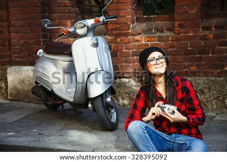 Smiling woman, wearing in red and black plaid shirt, jeans, glasses and hat, sitting on the pavement with retro camera, with brick building and silver scooter on a background, waist up