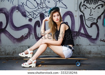 Beautiful young girl, with tattoo on her arm, wearing in cap, sandals, shirt and shorts, posing on her skateboard, near the wall with graffiti, in the park, full body